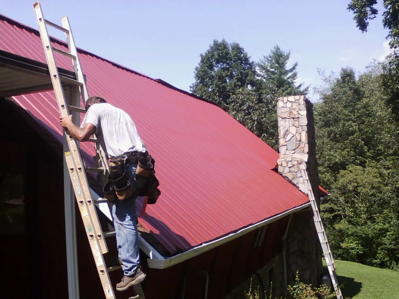 Roofer on a ladder beside a red metal roof and stone chimney — Soffit & Fascia Repair by Roofing Brothers in San Fernando